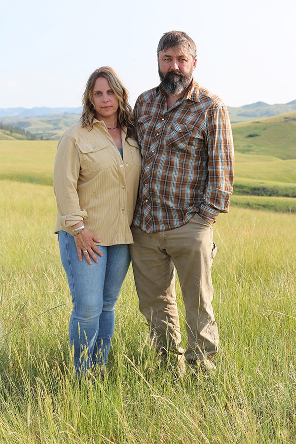 A woman and a man stand close together in a grassy field with rolling hills in the background, both wearing long-sleeved shirts and jeans, looking at the camera on a clear day.