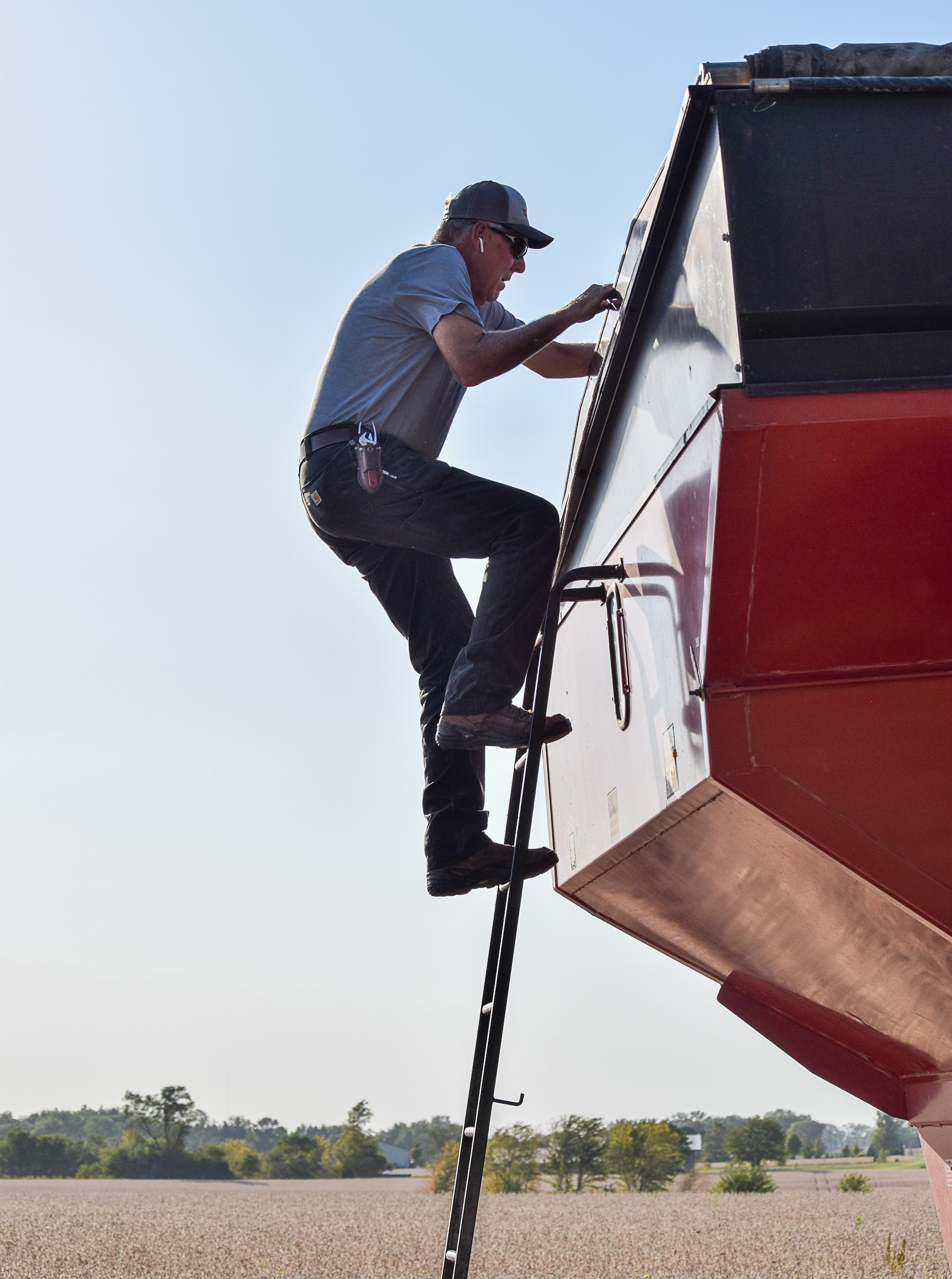 A person wearing a cap and sunglasses climbs a ladder attached to a large red agricultural machine in a field on a clear day. Trees and crops are visible in the background.