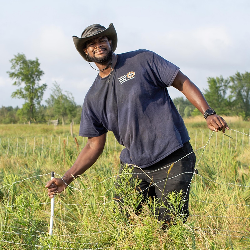 A man wearing a wide-brimmed hat and navy t-shirt works outdoors, adjusting a wire fence in a grassy field with trees in the background on a sunny day.