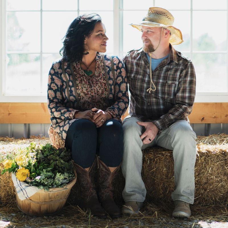 A woman and a man sit on a hay bale in a sunlit barn. The woman wears a patterned jacket and boots, and the man wears a straw hat and plaid shirt. A basket of fresh vegetables sits beside them. They are smiling at each other.